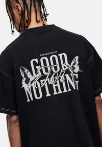 T-shirt noir oversize avec un imprimé graphique blanc de papillons et le texte "PRÉSENTÉ PAR GOOD FOR NOTHING" au dos.