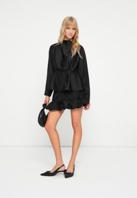 Černý saténový top s peplum a dlouhými rukávy, doplněný sukní s volánkem. Modelka drží černou kabelku a má na sobě odpovídající baleríny.