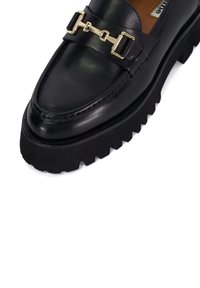 Zwarte leren loafers met een robuuste rubberen zool, voorzien van een goudkleurige decoratieve gesp en een glad, gepolijst oppervlak dat het strakke ontwerp benadrukt.