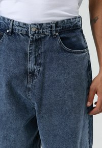 Donkere blauwe spijkerbroek met een losse pasvorm, voorzien van zakken aan de voorkant, een knoopsluiting en zichtbare stikseldetails.