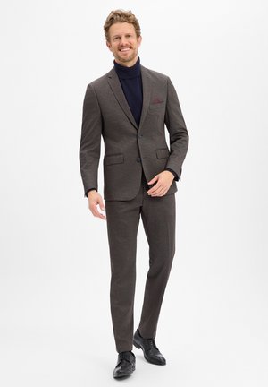 Glimlachende man die een donkerbruin geruit pak draagt met een colbert met twee knopen, een zwarte coltrui en zwarte nette schoenen, staand tegen een witte achtergrond.