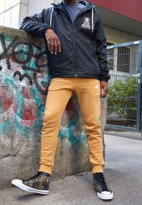 Černá lehká bunda s bílým logem, béžové tepláky a vzorované kotníkové tenisky. Oblečení je neformální a vhodné do městského prostředí.