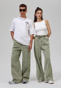 Grønne bukser med bred ben og struktureret overflade, parret med en hvid tanktop og en oversized hvid t-shirt med et grafisk hjerte-design.