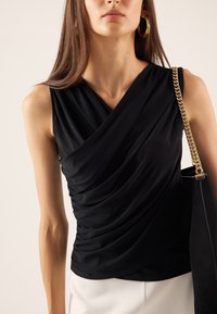 Top de color negro sin mangas con tejido drapeado, que presenta un diseño cruzado y una textura suave. En la imagen se puede ver un bolso con cadena dorada.