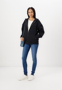 Moletom preto com fecho e grandes bolsos frontais, usado sobre uma blusa branca, combinado com jeans skinny azuis e ténis brancos. Pequena mala de ombro azul.