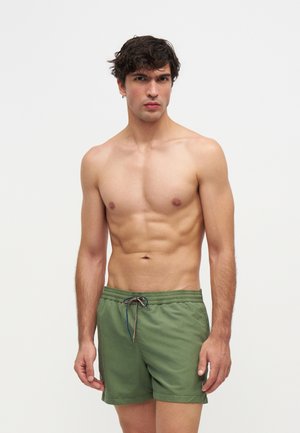Jeune homme aux cheveux foncés portant un short de bain vert, debout devant un fond clair uni, regardant droit devant lui.