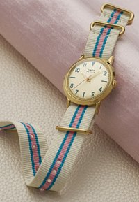 Montre Timex Marlin en or avec cadran crème, chiffres bleus, et bracelet en tissu blanc avec rayures bleues et roses, reposant sur une surface texturée.
