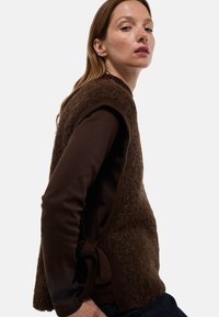 Gilet en laine marron avec des côtés ouverts et un accent noué. Finition texturée, bords côtelés, superposé sur un haut à manches longues marron foncé.