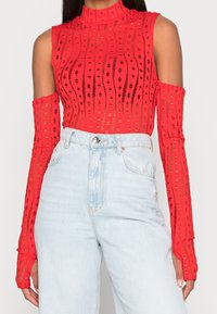 Red long-sleeve top with a high collar and cut-out design, featuring a ribbed texture and open shoulders, paired with light blue high-waisted jeans.
