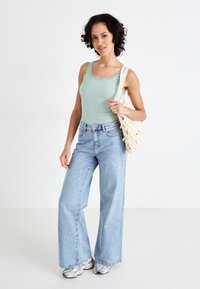 Canotta a costine verde chiaro con bordo in pizzo, abbinata a jeans ampi blu chiaro e sneakers grigie, mentre tiene una borsa in crochet color crema.
