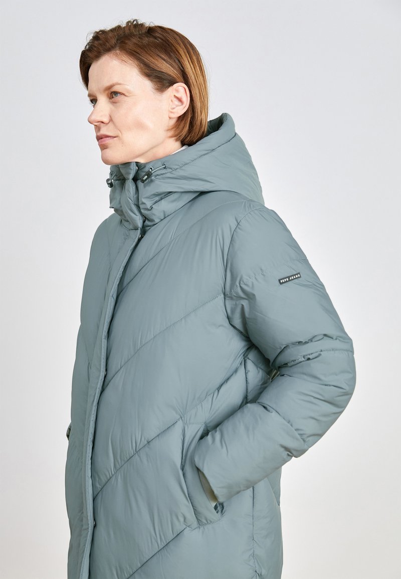 Abrigo acolchado en suave teal apagado, con un cuello alto, capucha, patrón de acolchado diagonal y dos bolsillos laterales. Material ligero con un acabado suave.