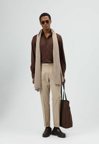 Bruine overhemd met knopen, beige broek, lichtbruine sjaal, bruine draagtas en donkere schoenen. Soepele stoffen, op maat gesneden pasvorm, benadrukt door neutrale tinten.