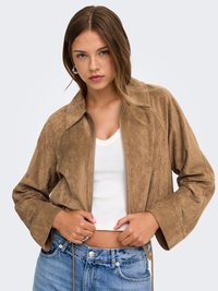 Jeune femme en veste en daim beige portée sur un crop top blanc et un jean bleu ajustant les cordons de sa veste sur un fond uni.