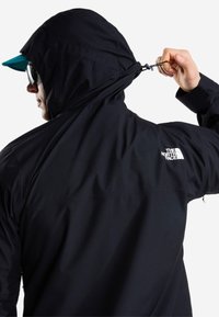Άτομο που φοράει μαύρο μπουφάν North Face τραβάει με το χέρι το κορδόνι της κουκούλας, φοράει γυαλιά ηλίου και πράσινο καπέλο κάτω από την κουκούλα, εμφανίζεται από πίσω.