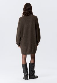 Üle suuruse pruuni kootud kampsunisärk pikkade varrukate, soonikkoes varrukaäärte ja ümarate äärtega. Sobitatud mustade põlveni ulatuvate kummikutsega.