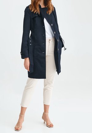 Woman wearing a navy buttoned trench coat over white shirt, beige cropped pants, and beige heeled sandals, standing against a light background.