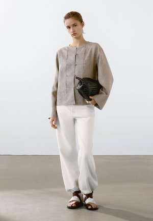 Haut gris en lin à manches longues avec détail de boutons sur le devant, associé à un pantalon blanc ample et des sandales brunes à lanières. Sac noir en tissage en accent.