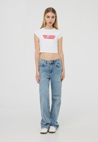 T-shirt bianca corta con testo rosso, abbinata a jeans azzurri. Il modello indossa sneakers bianche. Outfit semplice e casual con una vestibilità rilassata.