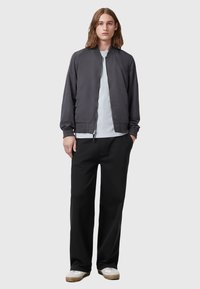 Szara kurtka bombowa o gładkiej powierzchni, z zapięciem na zamek, ściągaczami przy mankietach, nałożona na jasnoniebieską koszulę; w zestawie z czarnymi spodniami i trampkami.