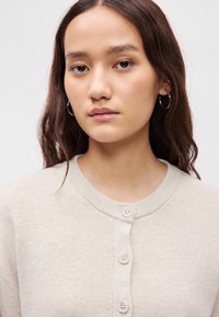 Ανοιχτό μπεζ ζακέτα με υφή πλέξης, featuring ριμπ λαιμόκοψη και τέσσερα κουμπιά μπροστά. Απλός σχεδιασμός με χαλαρή κομψότητα.