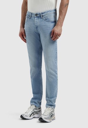 Man met lichtblauwe slim-fit jeans, witte sneakers en een wit T-shirt, staand tegen een effen lichte achtergrond.