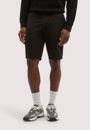 Mann trägt schwarze Shorts, weiße Sportsocken und graue-weiße Sportschuhe, steht vor einem einfarbigen Hintergrund.