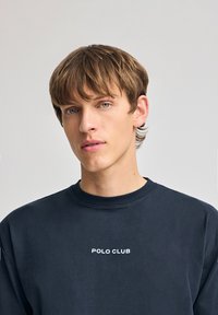Marinblå sweatshirt med rund halsringning och ribbade ärmslut, med "POLO CLUB" broderat i vitt på bröstet. Mjuk bomullstextur.