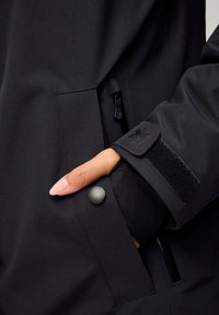 Zwarte waterdichte jas met een ritszak, drukknopen sluiting en een klittenband manchetten. Strakke textuur met minimalistische ontwerpdetails.