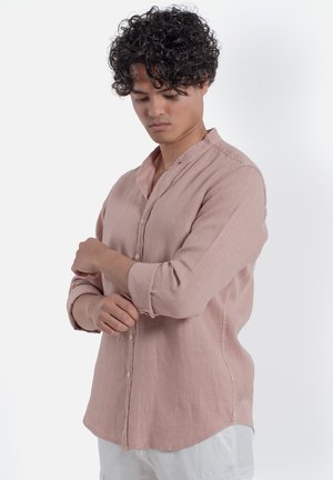 Giovane uomo con capelli ricci che si rimbocca la manica di una camicia rosa chiaro con bottoni, in piedi davanti a uno sfondo bianco semplice.
