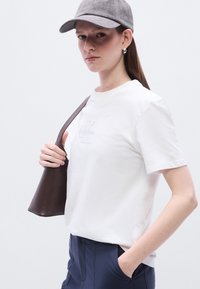 Λευκό βαμβακερό μπλουζάκι με κοντά μανίκια και διακριτικό κεντημένο λογότυπο. Γκρι καπέλο από ύφασμα και καφέ δερμάτινη τσάντα συμπληρώνουν την εμφάνιση.