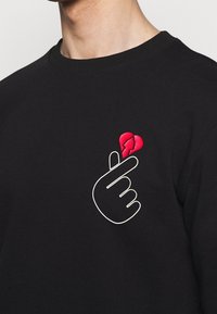 Sudadera negra con un diseño bordado de un gesto de mano blanca y un corazón rojo agrietado en el lado izquierdo del pecho.