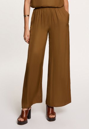 Mujer que lleva pantalones anchos de tiro alto sueltos de color marrón con bolsillos laterales y sandalias marrones de plataforma con tacón, de pie con una mano en el bolsillo.