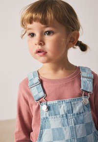 Lyserød langærmet skjorte kombineret med blå ternede overalls med sølvfarvede spænder, glat denimtekstur og kontrasterende mønstre.