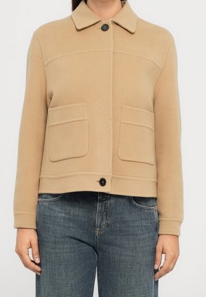 Beżowa wełniana kurtka z ostrym kołnierzem, dwoma przednimi kieszeniami naszywanymi oraz czarnymi zapięciami na guziki, noszona z niebieskimi jeansami.