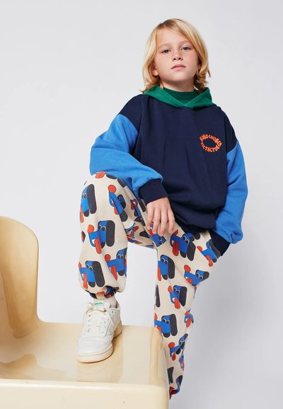 Marineblauer und himmelblauer übergroßer Kapuzenpullover mit grüner Kapuze, kombiniert mit hellbraunen Hosen, die bunte Cartoon-Hundemuster aufweisen. Weiße Sneaker.
