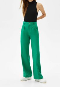 Gröna vida byxor med hög midja, med veck, knappstängning och en slät yta, kombinerat med vita sneakers.