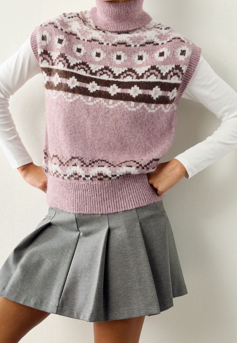 Femme portant un col roulé sans manches en tricot à motif rose sur un haut blanc à manches longues et une jupe plissée grise, les mains sur les hanches.