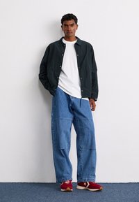 Veste noire légère, t-shirt blanc, pantalon en denim bleu ample et baskets rouges avec accents blancs et semelles marron.