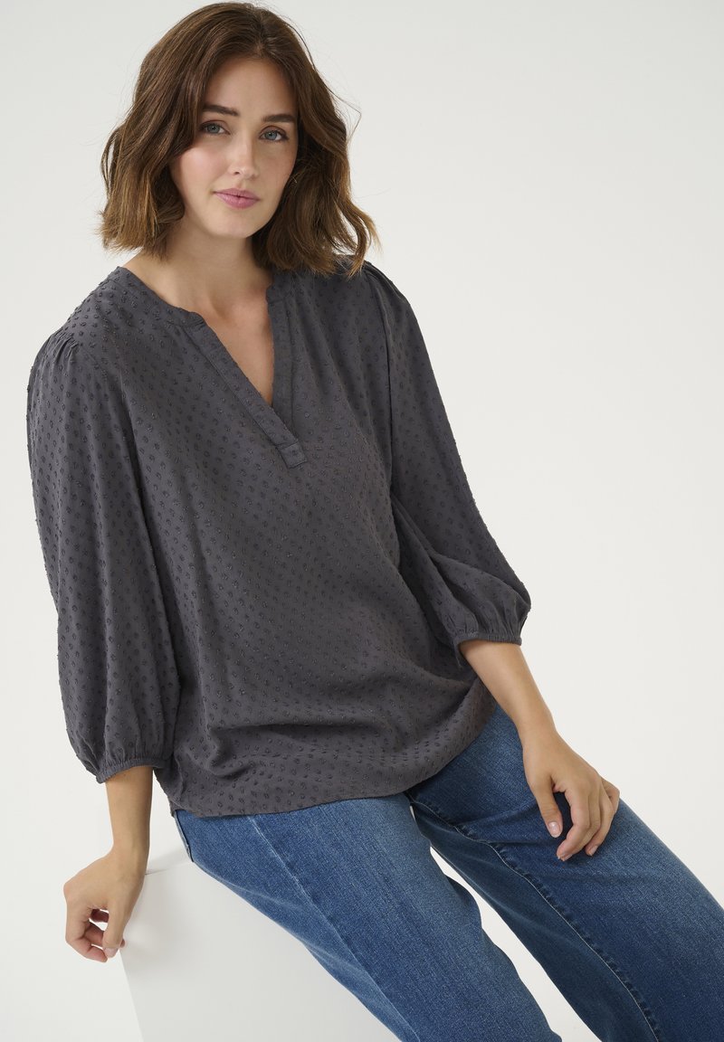 Donkergrijze blouse met pofmouwen en een V-hals, voorzien van textuurpatronen en een ontspannen pasvorm. In combinatie met blauwe denim jeans.