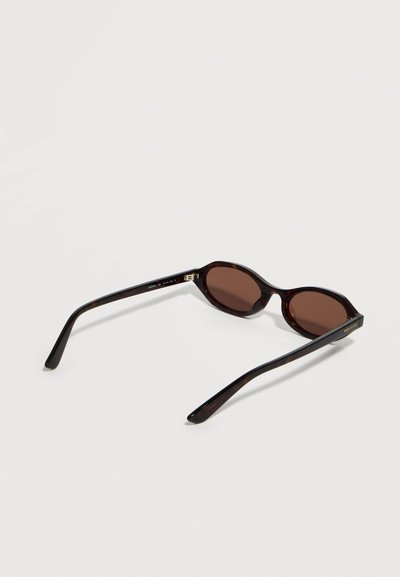 Οβάλ γυαλιά ηλίου με μοτίβο χελώνας, φακούς σκούρου καφέ και λεπτά μπράτσα, ανοιχτά τοποθετημένα σε ανοιχτή επιφάνεια.