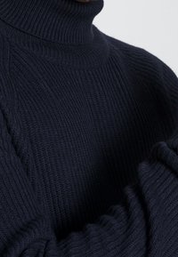 Marinblå ribbad stickad turtleneck-tröja med långa ärmar, visad från mitten av bröstet till nacken mot en neutral bakgrund.
