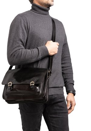 Homme portant un col roulé gris et une sacoche en cuir noir avec deux sangles à boucle sur l'épaule, debout devant un fond blanc.