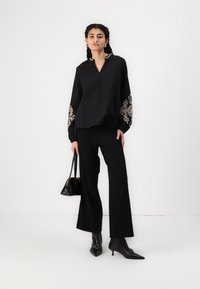 Blusa nera a maniche lunghe con intricate ricami beige sulle maniche, abbinata a pantaloni neri a gamba larga e stivaletti con tacco. Accessorio borsa nera.