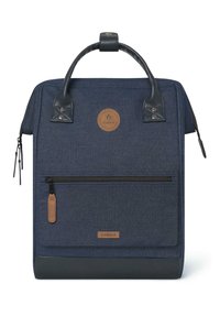 Sac à dos en tissu bleu marine avec des accents en cuir, une poignée supérieure, une poche zippée à l'avant et un écusson de logo circulaire. Texture lisse, forme rectangulaire.