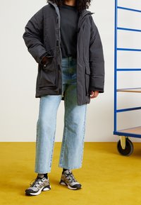 Vadderad svart parkas med huva och fickor, i kombination med ljusblå vida jeans och grå sportskor med svarta accenter.