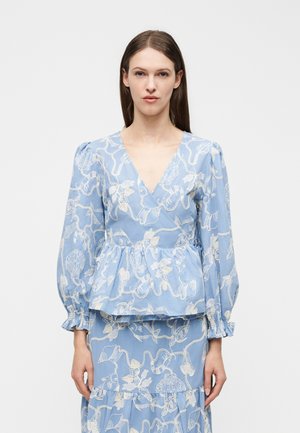 Woman facing forward wearing a light blue V-neck top and skirt with white seashell and chain pattern, long puffed sleeves, and ruffled cuffs.