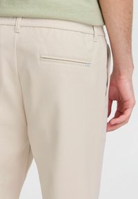 Beige pants with a back welt pocket and belt loops worn by a person in a light green shirt, arm visible at the side.