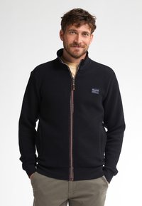Giacca nera con zip, caratterizzata da un motivo testurizzato, colletto alto, tasche laterali e zip a contrasto. Panno con logo sul lato sinistro del petto.