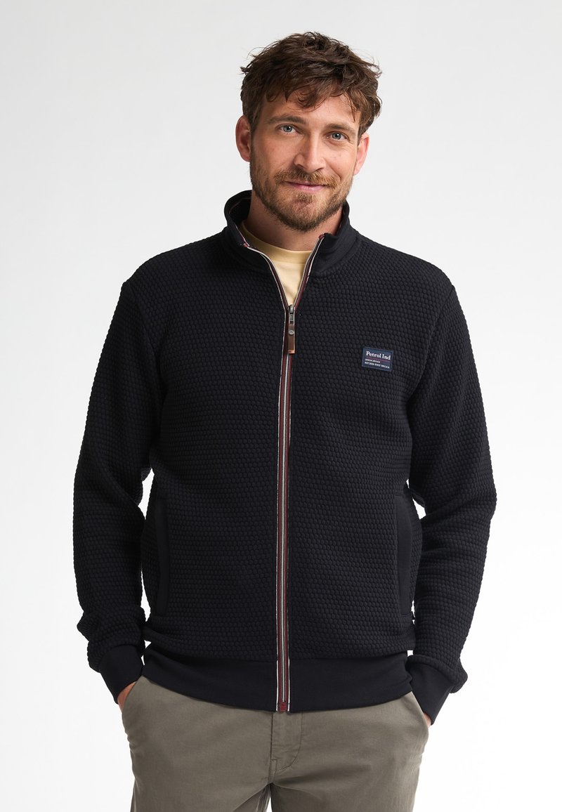 Svart zip-up jacka med texturerat mönster, hög krage, sidofickor och kontrasterande dragkedja. Logopatch på vänster bröst.