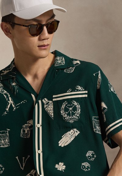 Mężczyzna w ciemnozielonej koszulce z krótkim rękawem z białymi sportowymi nadrukami, brązowych okularach przeciwsłonecznych i białej czapce, patrzący w lewo na neutralnym tle.